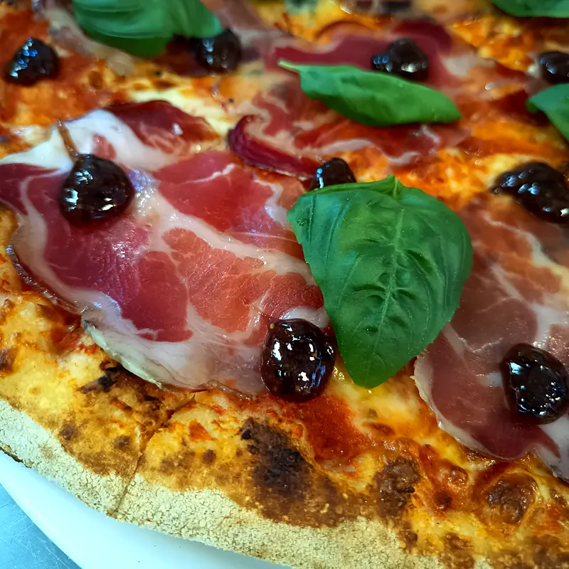 Gros plan d'une pizza tomate garnie de jambon cru, d'olives noires et de feuilles de basilic frais