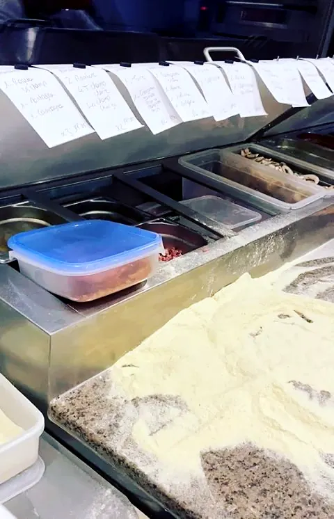Plan de travail du pizzaiolo avec bacs d'ingrédients réfrigérés, bons de commande et farine sur le granit